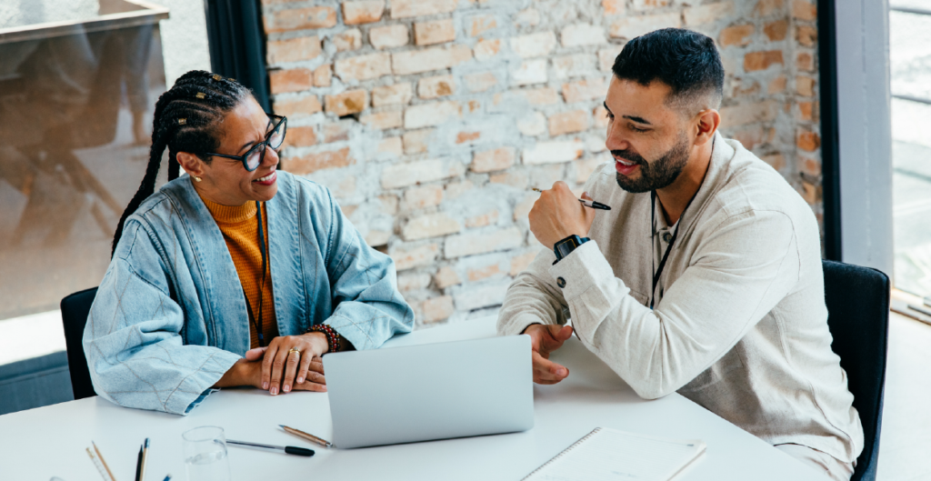 Two colleagues in conversation at a table with a laptop, exchanging ideas during a workplace mentorship discussion.