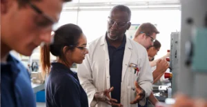 An instructor in lab coat explains a concept to students wearing safety glasses in a hands-on training workshop, with equipment and workstations visible in the background